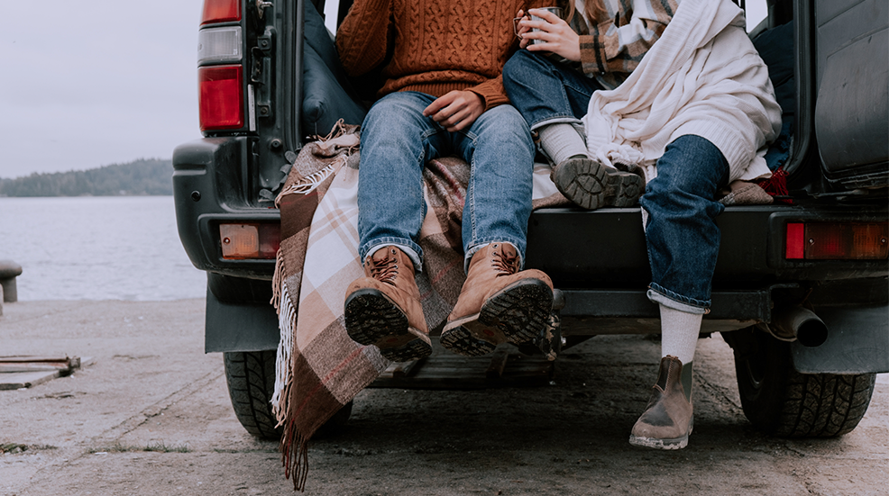 A couple sat in the boot of their car, drinking from travel mugs and dressed in autumnal clothing